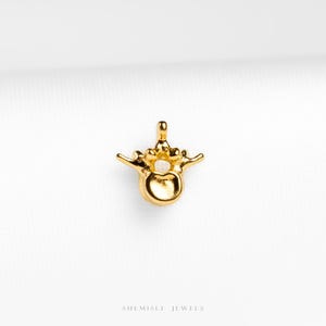 May include: A gold-colored pendant shaped like a human vertebra, featuring a central circular opening and lateral projections. The piece has a smooth, polished metallic finish and includes the text SHEMISLI JEWELS at the bottom.