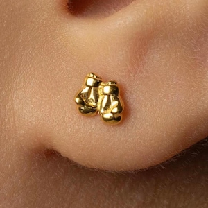 May include: A small, shiny gold-toned stud earring shaped like a pair of miniature boxing gloves. The piece features a polished metallic finish with detailed indentations representing the laces and padding of the gloves, secured to the earlobe.