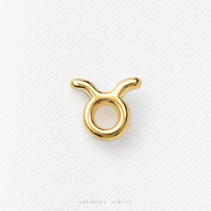 May include: A gold-toned metal charm shaped like the Taurus zodiac symbol, featuring a circular center with two curved horns extending upward. The piece has a smooth, polished finish and is centered above the text SHEMISLI JEWELS.