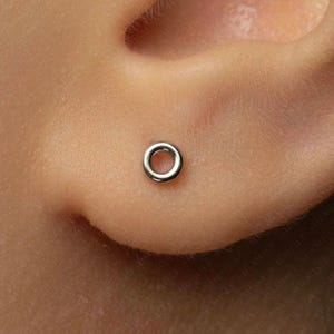 May include: A small, circular, silver-toned metal hoop earring positioned in an earlobe. The earring features a simple, smooth, polished finish with a hollow center, creating a minimalist ring shape that sits flush against the skin of the ear.