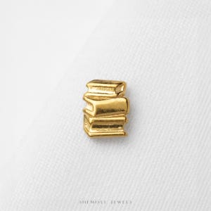 May include: A small gold-toned charm shaped like a stack of three books. The charm features a polished metallic finish with defined horizontal lines representing the pages and covers of the books. The text SHEMISLI JEWELS is printed below.