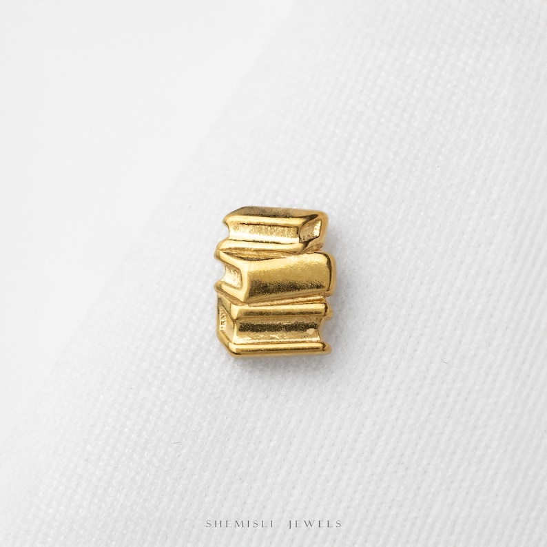 May include: A small gold-toned charm shaped like a stack of three books. The charm features a polished metallic finish with defined horizontal lines representing the pages and covers of the books. The text SHEMISLI JEWELS is printed below.
