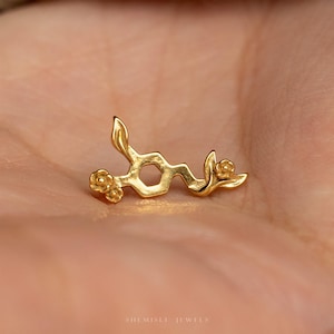 May include: A gold-toned pin shaped like a chemical molecular structure, featuring a central hexagonal ring connected to linear segments. The design is accented with small, sculpted floral motifs and leaf-like shapes at the ends of the structure.