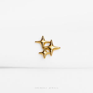 May include: A gold-toned metal stud earring featuring a cluster of three four-pointed star shapes of varying sizes. The surface is smooth and reflective, catching the light. The text "SHEMISLI JEWELS" is printed in small, dark letters at the bottom.