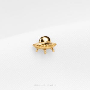 May include: A small, gold-colored UFO charm with a domed top and three small legs. The charm is highly detailed and appears to be made of metal. The background is a plain white surface. The text "SHEMISLI JEWELS" is at the bottom.