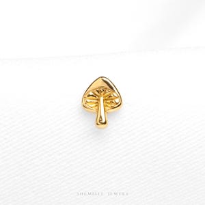 May include: A small gold-colored mushroom stud earring with a polished finish. The cap features detailed, etched radial lines radiating from the center stem. The text "SHEMISLI JEWELS" is printed in small, dark letters at the bottom of the frame.