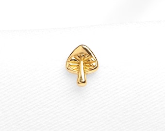 Op de afbeelding: Een kleine goudkleurige paddenstoel oorsteker met een gepolijste afwerking. De hoed heeft gedetailleerde, gegraveerde radiale lijnen die vanuit de centrale steel uitstralen. De tekst "SHEMISLI JEWELS" staat in kleine letters onderaan.