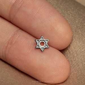 May include: A small, silver-toned metal stud earring shaped like a six-pointed Star of David. The design features a central hexagonal cutout surrounded by a geometric frame, creating a clean, open-work appearance. The piece is polished and metallic.