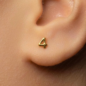 May include: A small, polished gold-tone stud earring shaped like the numeral four. The earring features a smooth, rounded surface and is positioned directly on the skin of an earlobe, showcasing a simple, minimalist design with a metallic finish.