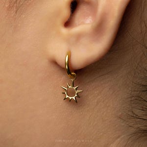 May include: A gold-toned hoop earring featuring a small, dangling sun-shaped charm with radiating points. The earring is secured through the earlobe. The text SHEMISLI JEWELS is printed in small, light-colored letters at the bottom of the image.