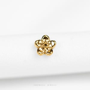 May include: A small, gold-colored flower-shaped jewelry piece. The jewelry has five rounded petals and a detailed center. The piece is set against a white background. The text "SHEMISLI JEWELS" is at the bottom.