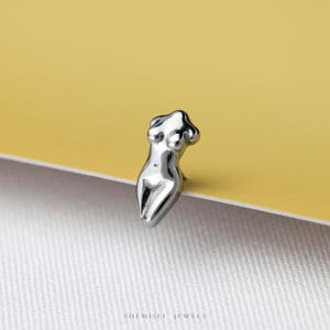 May include: A silver-toned metal stud earring shaped like a stylized human torso. The piece features a smooth, reflective surface with defined curves representing the chest and waist. The text SHEMISLI JEWELS is printed at the bottom of the image.