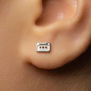 May include: A small, silver-toned stud earring shaped like a rectangular cassette tape. The earring features a metallic finish with engraved details representing the two circular spools and the central label area of a classic audio cassette.