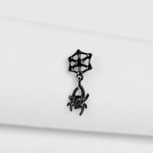 May include: A black earring featuring a spider web design at the top, connected to a dangling spider charm. The spider has eight legs and a detailed body. The earring is set against a white background.