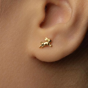 May include: A gold-colored stud earring in the shape of a running rabbit is shown on an ear. The earring is small and detailed, with visible ears, legs, and a tail. The earring is set against the skin of the ear.