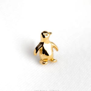 May include: A small, polished gold-toned stud earring shaped like a standing penguin. The piece features a smooth, reflective surface with a defined beak, head, and flippers, capturing the distinct silhouette of the bird in a minimalist, metallic form.