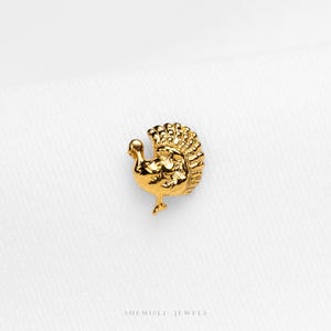 May include: A small gold-toned metal stud earring shaped like a turkey with a detailed fan-shaped tail and textured body. The piece features a polished finish and is positioned above the text SHEMISLI JEWELS printed in a simple black font.