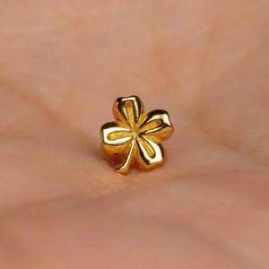 May include: A small, polished gold-toned stud earring shaped like a four-leaf clover. The piece features a smooth, reflective surface with defined indentations separating each of the four rounded petals, creating a symmetrical floral design.
