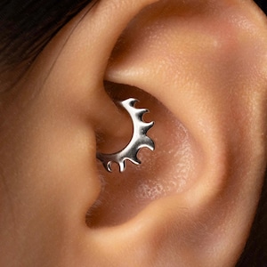May include: A silver-toned circular daith piercing ring featuring a series of uniform, pointed, gear-like teeth along the outer edge. The metallic jewelry is positioned within the inner cartilage fold of the ear, showcasing a polished finish.