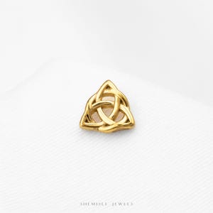 May include: A small, polished gold-tone stud earring featuring a triangular triquetra knot design with three interlocking loops. The piece has a smooth, reflective metallic finish and is marked with the text SHEMISLI JEWELS at the bottom.