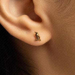 May include: A small, polished gold stud earring shaped like a bird, featuring a smooth, reflective surface and detailed contours that define the head, wings, and tail of the avian figure, securely positioned on the lower part of an earlobe.