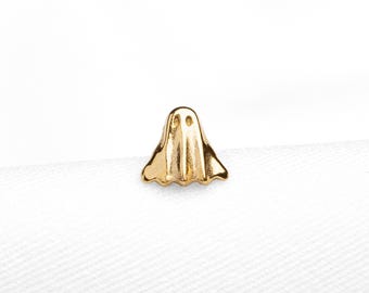 Könnte beinhalten: Ein kleiner goldfarbener Ohrstecker in Form eines Geistes mit zwei kreisförmigen Aussparungen für die Augen und einem gewellten unteren Rand. Das Stück hat eine polierte metallische Oberfläche auf weißem Stoff mit dem Text SHEMISET JEWELS.