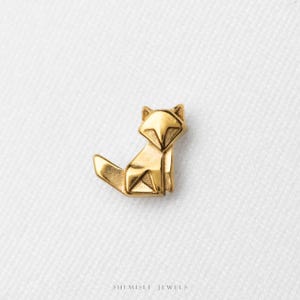 May include: A gold-toned fox-shaped charm featuring geometric, origami-inspired facets and sharp, angular lines. The small, metallic fox is depicted in a seated position with its tail extended. The text SHEMISLI JEWELS is printed below.