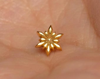 May include: A small, metallic gold-toned stud earring shaped like a six-pointed snowflake. The design features faceted, pointed petals radiating from a central point, creating a reflective, star-like appearance with a polished, smooth finish.
