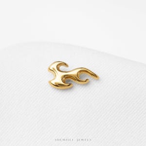 May include: A polished gold-tone metal earring shaped like a stylized flame with smooth, curved edges. The piece features a reflective surface and a tapered design. Below the earring, the text SHEMISLI JEWELS is printed in a small, dark font.