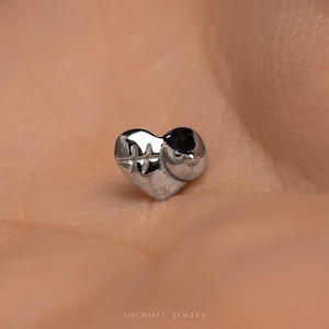 May include: A small, silver heart-shaped stud earring. The earring has a textured surface with a cross-like design on the front. The earring is shown against a warm, neutral background. The text "SHI MISLI JEWELS" is at the bottom.