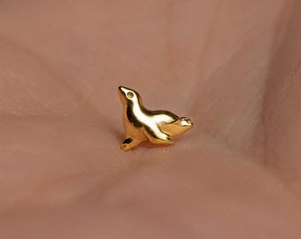 May include: A small, polished gold-tone stud earring shaped like a sea lion in a swimming pose. The piece features a smooth, reflective surface with a simple, minimalist silhouette and a small circular indentation representing the eye of the animal.