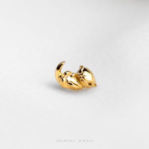 May include: A small, polished gold charm shaped like a sea otter lying on its back. The metallic figure features a smooth, reflective surface with subtle indentations defining the head, paws, and curled tail. The text SHEMISLI JEWELS is visible.