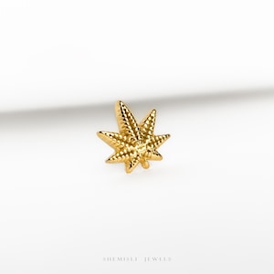 May include: A gold-toned stud earring shaped like a seven-pointed leaf with a textured, dimpled surface. The jewelry piece features a central stem and distinct serrated edges, reflecting light across its metallic finish. The text SHEMISLI JEWELS is visible.