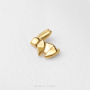 May include: A gold-toned stud earring shaped like a stylized rabbit in a geometric, origami-inspired design. The piece features a polished, reflective surface with distinct angular facets and clean lines. The text SHEMISLI JEWELS is printed below.