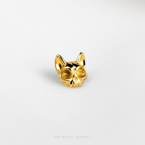 May include: A small, polished gold-tone stud earring shaped like a stylized cat skull with pointed ears and hollow eye sockets. The piece features a smooth, reflective metallic finish and is marked with the text SHEMISLI JEWELS at the bottom.