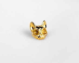 May include: A small, polished gold-tone stud earring shaped like a stylised cat skull with pointed ears and hollow eye sockets. The piece features a smooth, reflective metallic finish and is marked with the text SHEMISLI JEWELS at the bottom.
