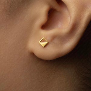 May include: A small, gold-colored stud earring with a geometric diamond shape. The earring is worn on an ear, showcasing its minimalist design and polished finish. The earring's design features a solid bottom half and an open top half.