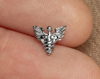 May include: A small, silver-toned metal pin or charm featuring a caduceus symbol. The design consists of a central staff with two wings extending from the top and a snake coiled around the staff, creating a detailed, polished medical emblem.