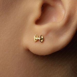 May include: A small, gold-colored bow-shaped stud earring. The earring is positioned on an ear, showcasing its delicate design and polished finish. The bow's details are clearly visible, highlighting its symmetrical shape and texture.