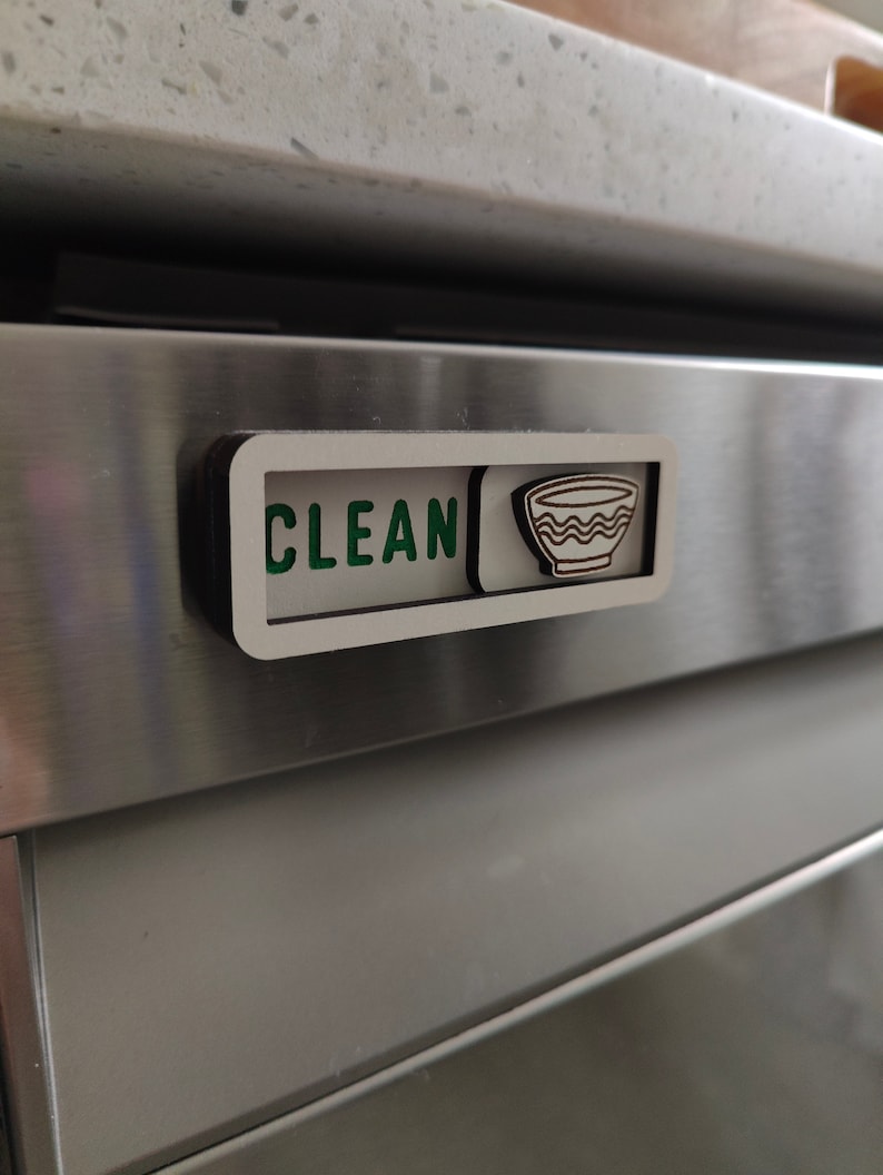 May include: A wooden dishwasher magnet with the word "CLEAN" in green letters and a stylized image of a bowl. The magnet is attached to a stainless steel dishwasher.