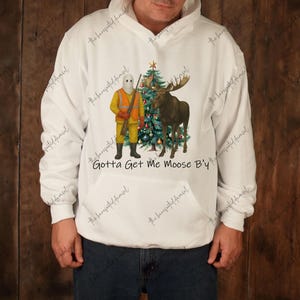Könnte beinhalten: Weißer Kapuzenpullover mit einem Aufdruck, der eine maskierte Figur in Jagdkleidung, einen Weihnachtsbaum und einen Elch zeigt. Der Text "Gotta Get Me Moose B'y" ist unter dem Motiv aufgedruckt.