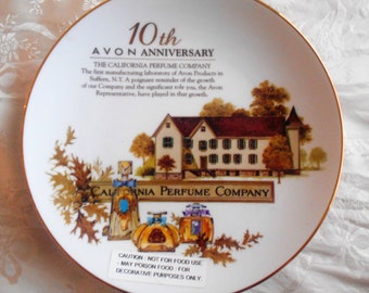 May include: A white ceramic plate with a gold rim, featuring a 10th anniversary illustration of the California Perfume Company. The plate depicts a building with a red roof and a sign that reads "California Perfume Company". The plate also features a text description of the company's history and a cautionary note that the plate is not for food use.