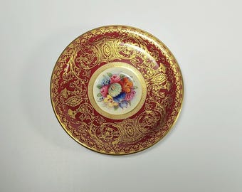 May include: A round porcelain saucer featuring a deep burgundy border decorated with intricate gold filigree patterns. The center of the saucer displays a circular white medallion containing a colorful bouquet of painted flowers in shades of pink, blue, and orange.