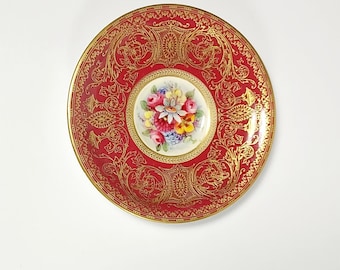 May include: A round porcelain saucer featuring a deep red border decorated with intricate gold filigree patterns. The center of the saucer displays a hand-painted floral arrangement with pink, yellow, and blue flowers on a white background.