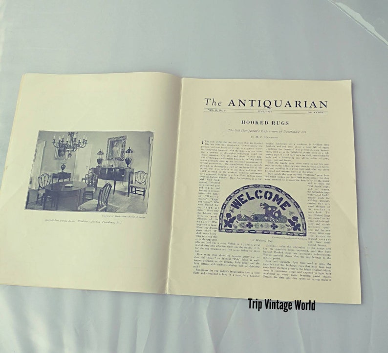 Op de afbeelding: Een vintage tijdschriftpagina met een artikel over gehaakte tapijten. Het artikel heeft de titel "Hooked Rugs: The Old Home-made Art: A Expression of Pioneer Art." De pagina bevat ook een zwart-witfoto van een eetkamer met een kroonluchter en een tapijt op de vloer. Het tapijt is een blauwe en witte welkomstmat met de woorden "Welcome" in het midden.