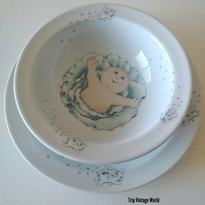 Può includere: Una ciotola in ceramica bianca con un design di cavolo blu e verde e una bambola di bambino all'interno. La ciotola è circondata da un piatto bianco con un design di cavolo blu e verde e una bambola di bambino all'interno. Il piatto è decorato con piccole stelle bianche e quattro coniglietti bianchi.