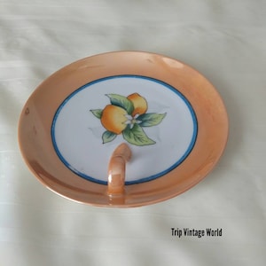 Puede incluir: Un plato de cerámica vintage con un borde color melocotón y un centro blanco con un diseño pintado de naranjas y hojas. El plato tiene un asa pequeña y curvada y un borde azul. El texto "Trip Vintage World" es visible.
