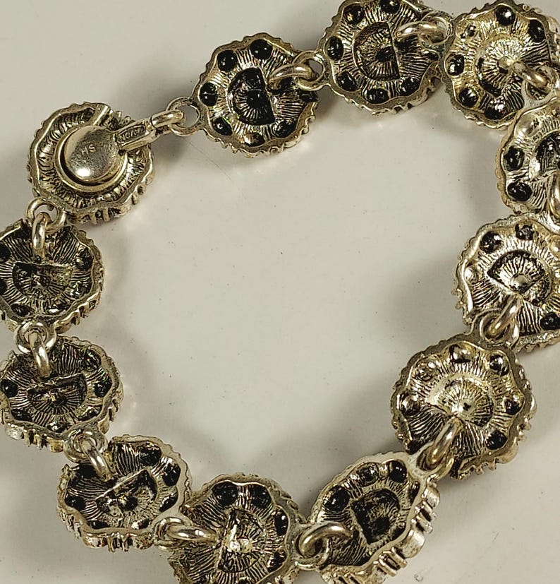May include: Silver bracelet with flower-shaped links and black detailing. The bracelet has a secure clasp and is intricately designed. The links are connected by small chains, creating a flexible and elegant accessory. The bracelet is a stylish piece of jewellery.