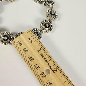 May include: Silver necklace with a repeating floral design, each element featuring a black center. The necklace is measured with a wooden ruler, showing the inch measurements. The necklace is on a white background.