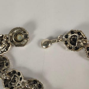 May include: Close-up of a silver necklace with a floral design. The necklace has small, circular links with black accents and a secure clasp. The clasp is connected to a series of decorative links.